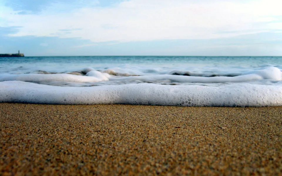 Close-up view of a foamy ocean wave receding from a sandy beach under a cloudy sky.