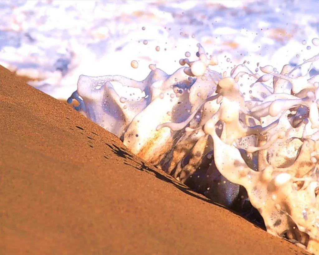 A close-up of a foamy ocean wave splashing onto a sandy beach, with water droplets suspended in the air.