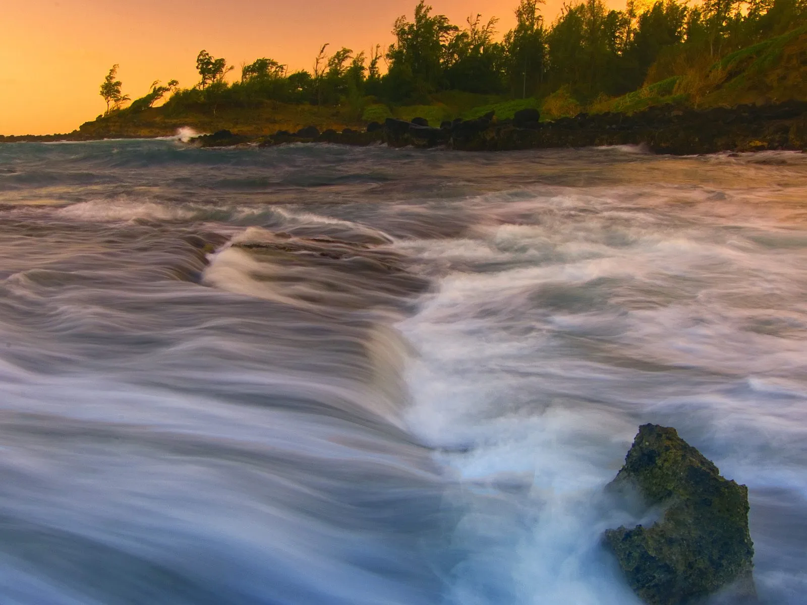 Long exposure of ocean waves flowing over rocks with a warm sunset sky in the background.