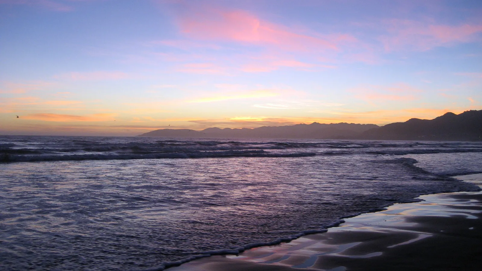A serene beach scene with gentle waves lapping the shore during a colorful sunset with mountains in the distance.