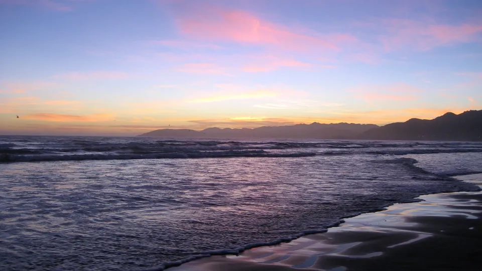 A serene beach scene with gentle waves lapping the shore during a colorful sunset with mountains in the distance.