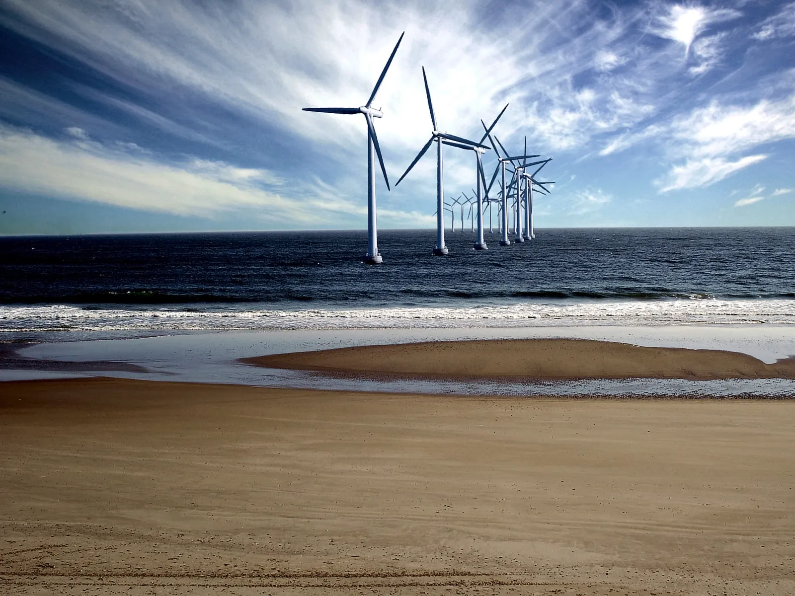 A line of wind turbines stands in the ocean near a sandy beach under a cloudy blue sky.