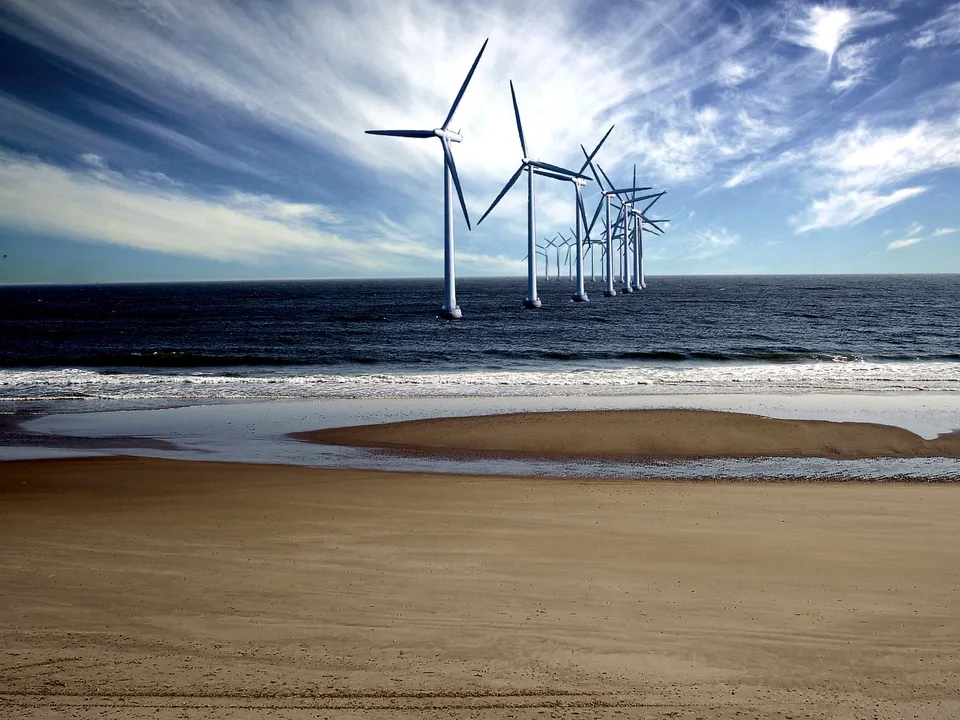 A line of wind turbines stands in the ocean near a sandy beach under a cloudy blue sky.