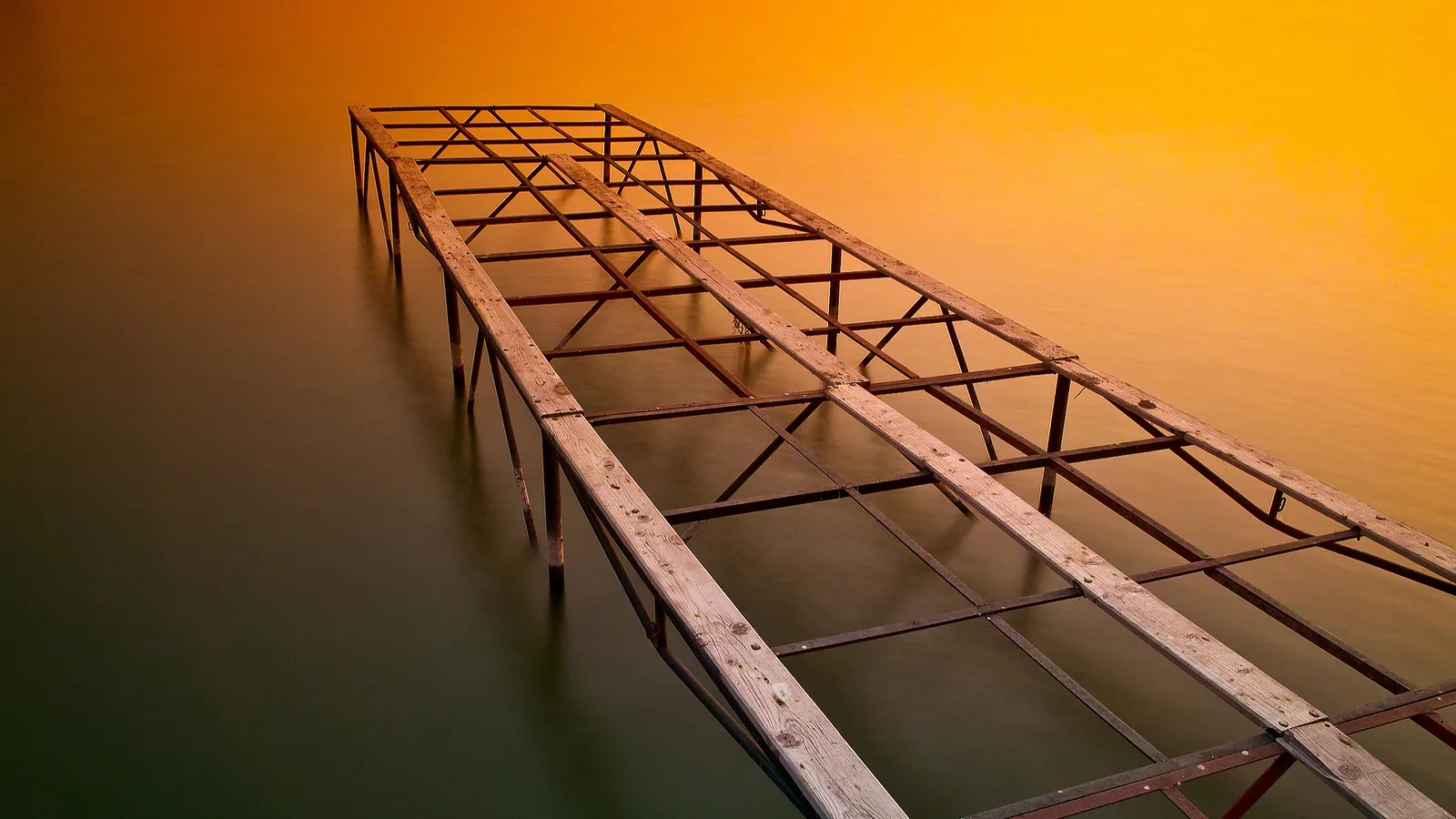 An old wooden and metal pier extends into dark, calm water under a vibrant orange sky.