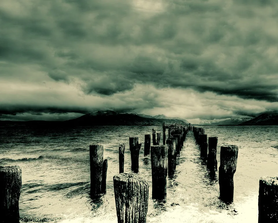 A line of weathered wooden posts extends into a choppy lake under a dramatic, cloudy sky.