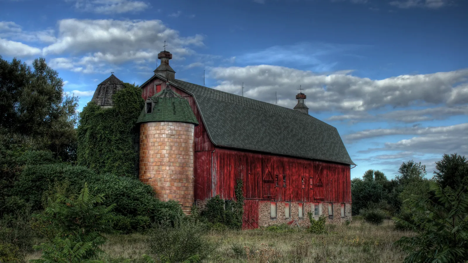 A weathered red barn with a silo covered in ivy stands in a field under a dramatic sky with clouds.