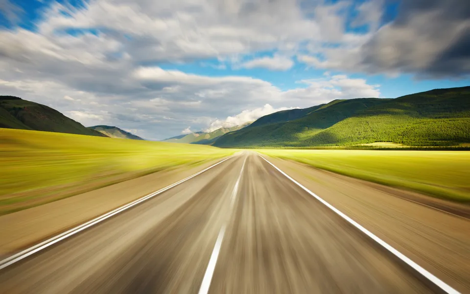 A blurred asphalt road stretches into the distance, flanked by vibrant green fields and rolling hills under a cloudy sky.