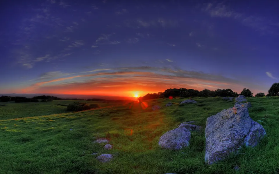 The sun sets on the horizon casting a warm orange glow on rolling green hills dotted with large rocks.
