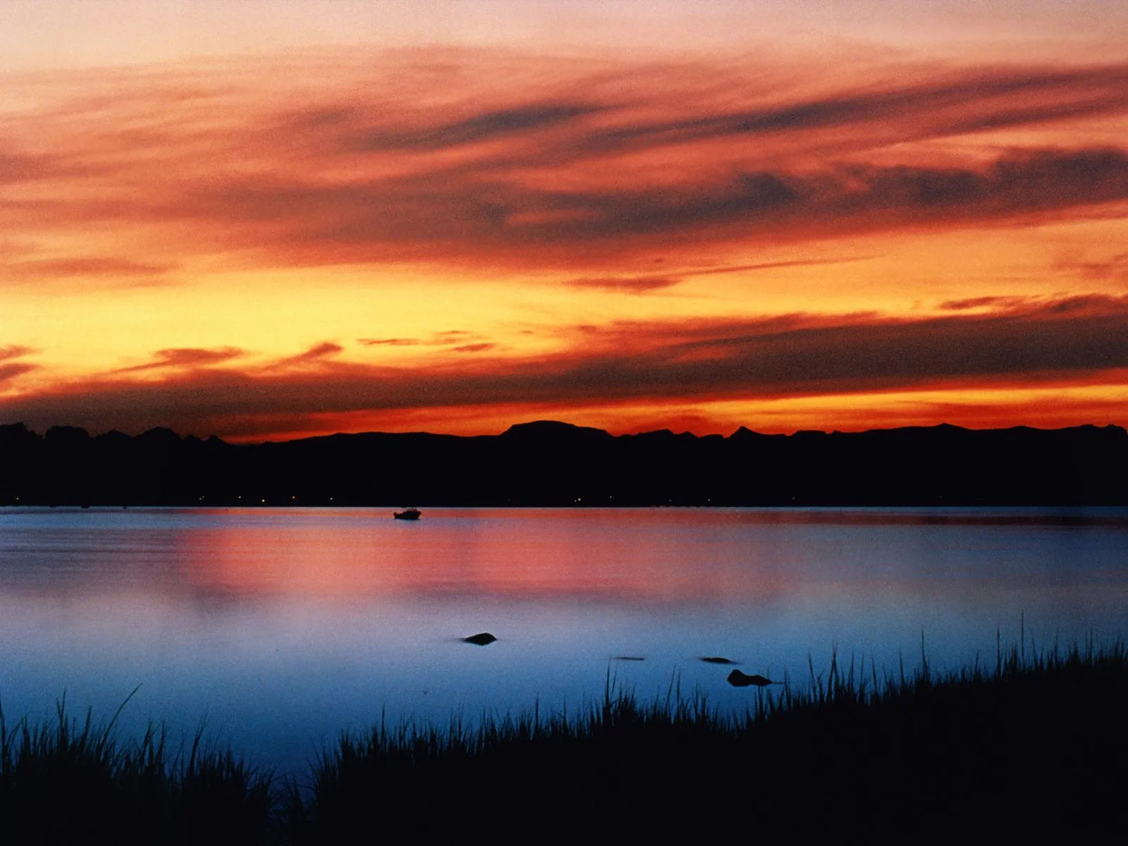 Fiery orange and yellow sunset reflecting on a calm lake with silhouetted mountains on the horizon.