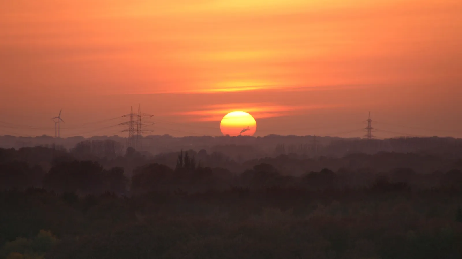 The sun sets behind a hazy, silhouetted landscape with power lines and wind turbines visible.