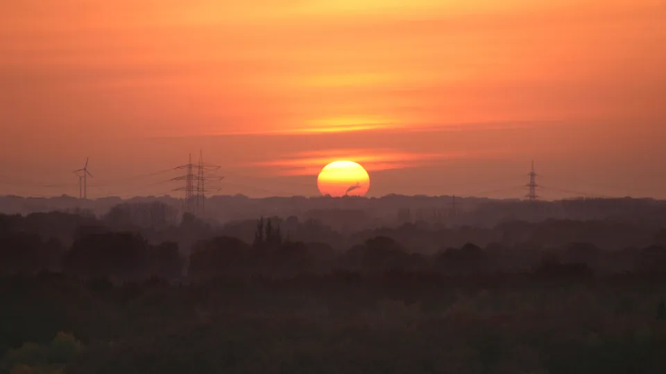 The sun sets behind a hazy, silhouetted landscape with power lines and wind turbines visible.