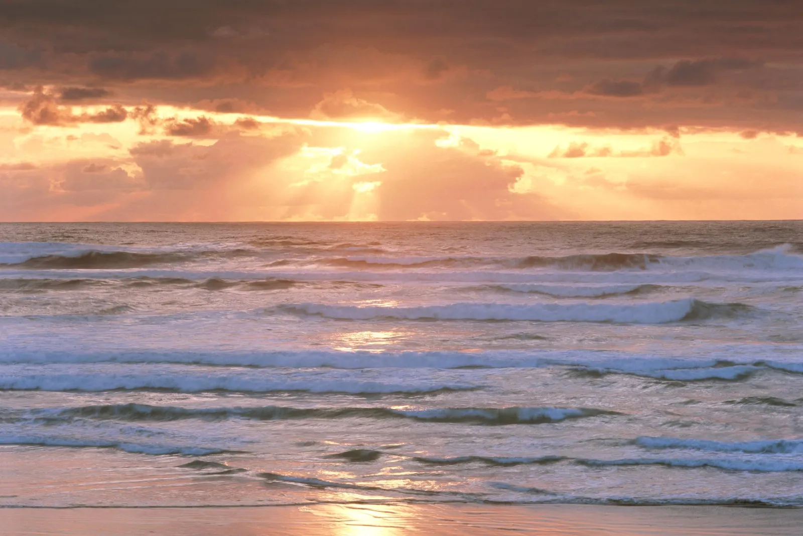 The sun sets over the ocean, casting golden rays through dramatic clouds onto the water.