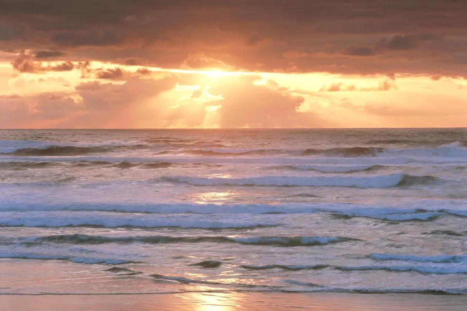 The sun sets over the ocean, casting golden rays through dramatic clouds onto the water.