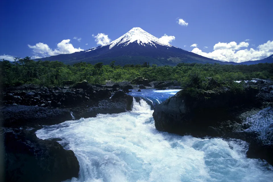 A snow-capped volcano looms over a powerful waterfall cascading over dark rocks.
