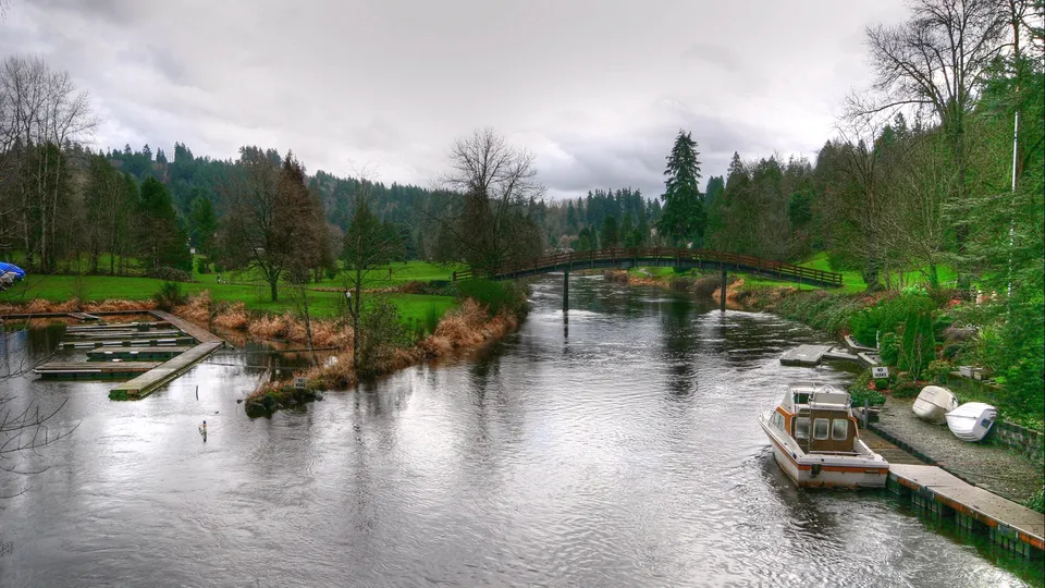 A river reflects an overcast sky, with a boat docked and a marina visible.