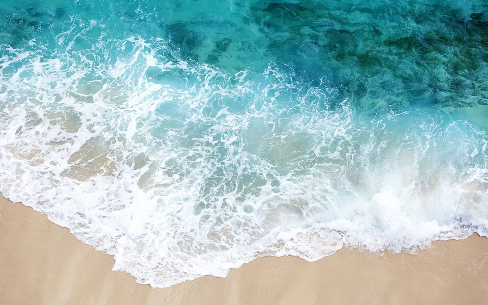 An overhead view of turquoise ocean waves crashing onto a sandy beach, creating white foam.