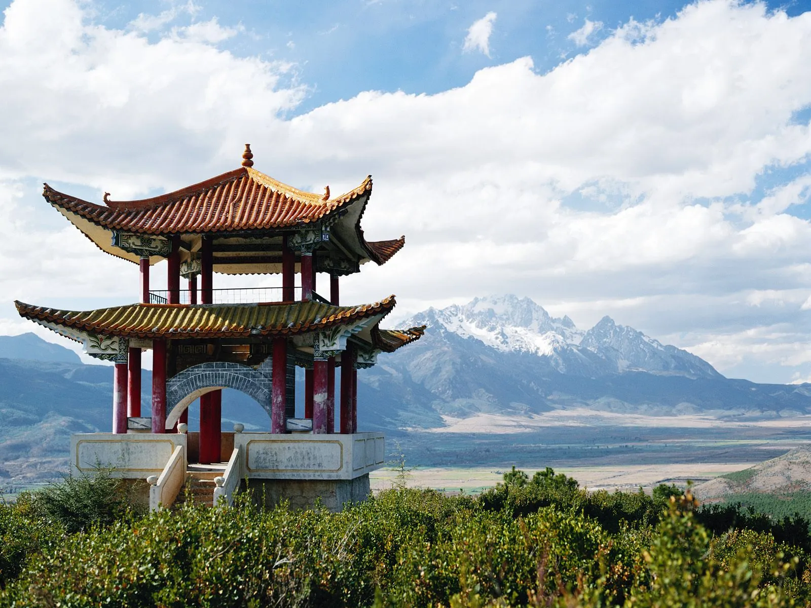 A traditional Chinese pagoda sits on a hill with snow-capped mountains in the background under a cloudy sky.
