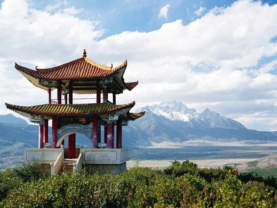 A traditional Chinese pagoda sits on a hill with snow-capped mountains in the background under a cloudy sky.