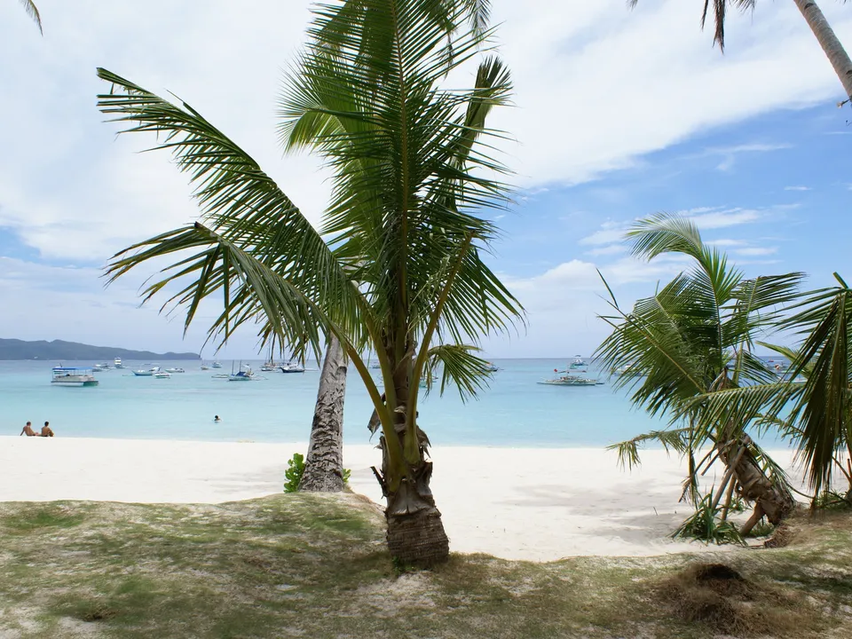 Palm trees overlook a white sand beach with calm turquoise waters where several boats are anchored offshore under a partly cloudy sky.