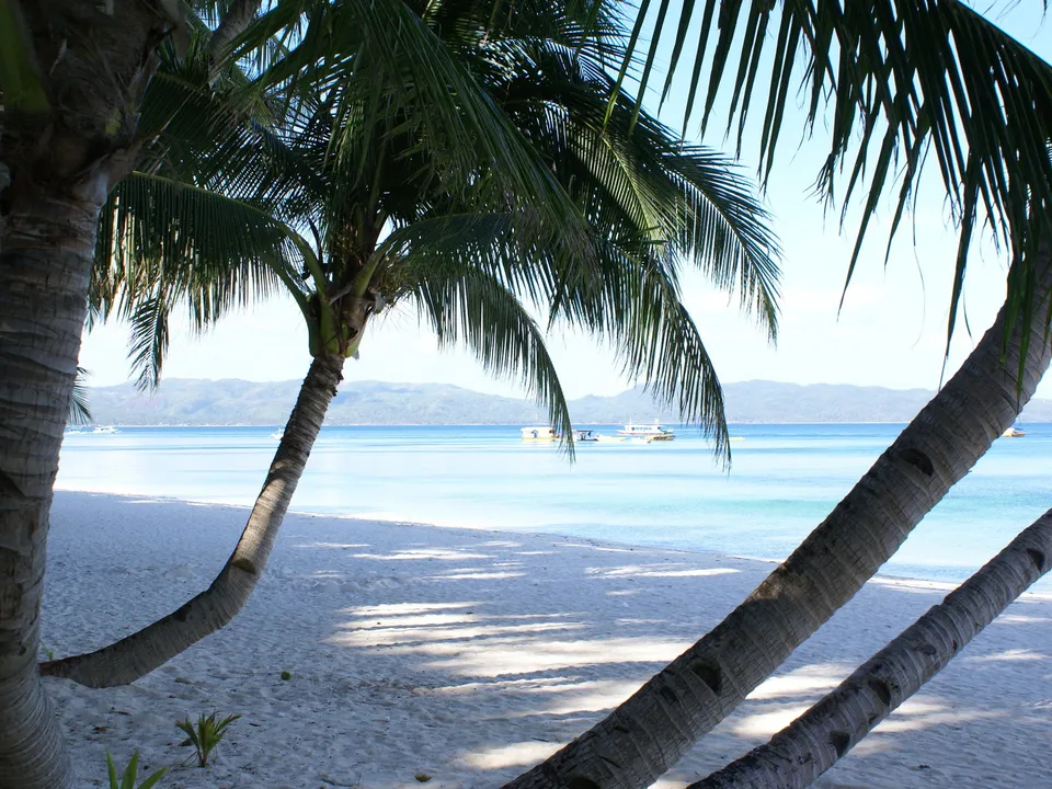 Palm trees frame a white sand beach leading to calm turquoise waters with distant islands and boats.