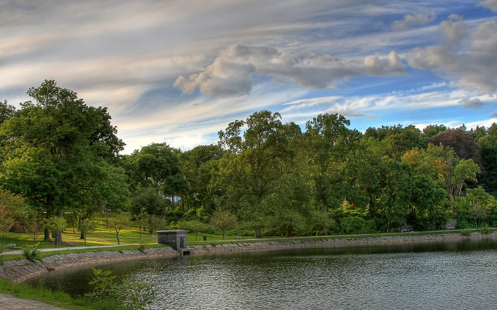A calm lake with rippling water is bordered by a stone embankment and a path, with a dense forest of green trees in the background under a cloudy sky.