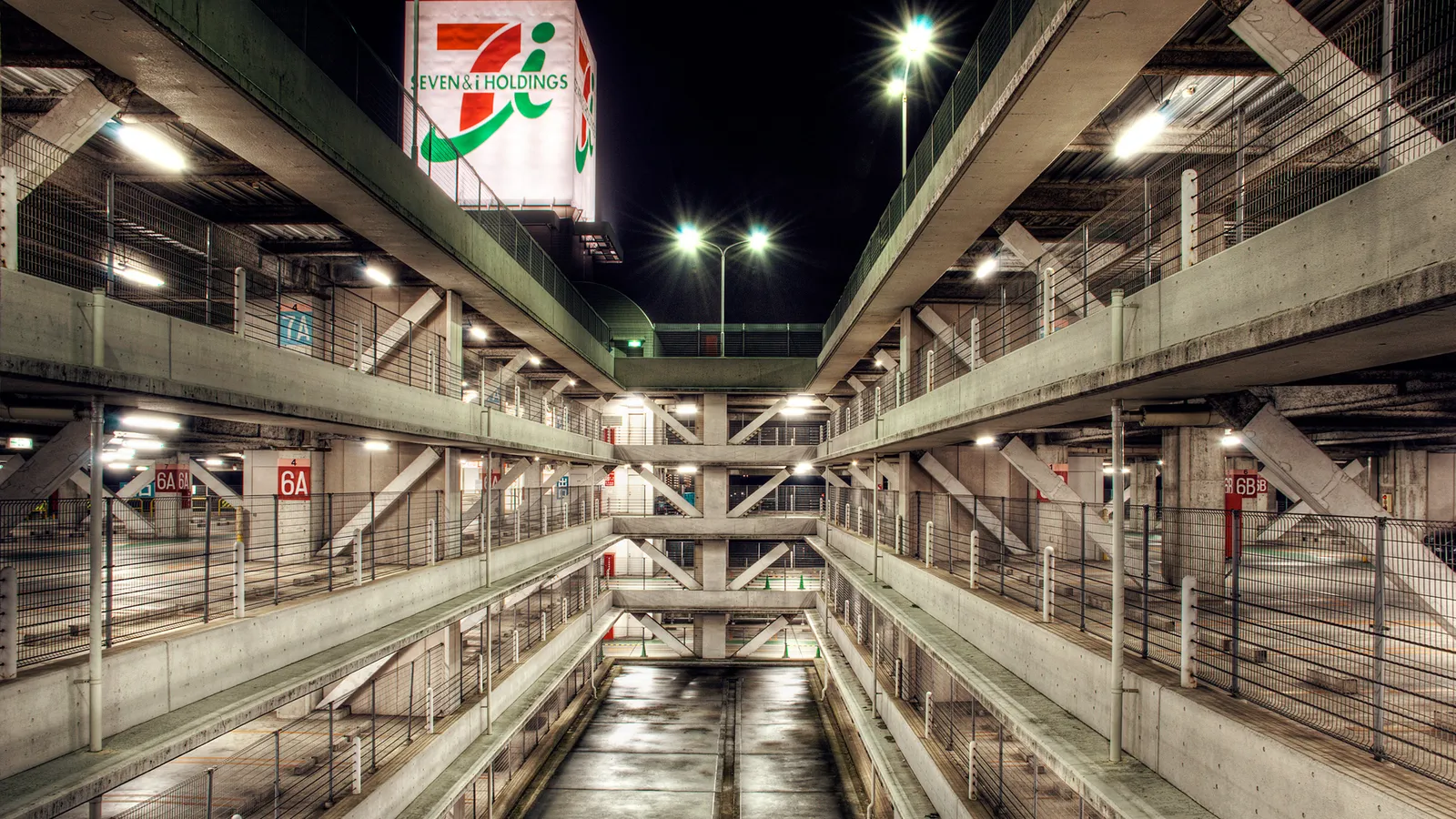 A multi-level parking garage at night, illuminated by artificial lights, with a large Seven & i Holdings sign visible.