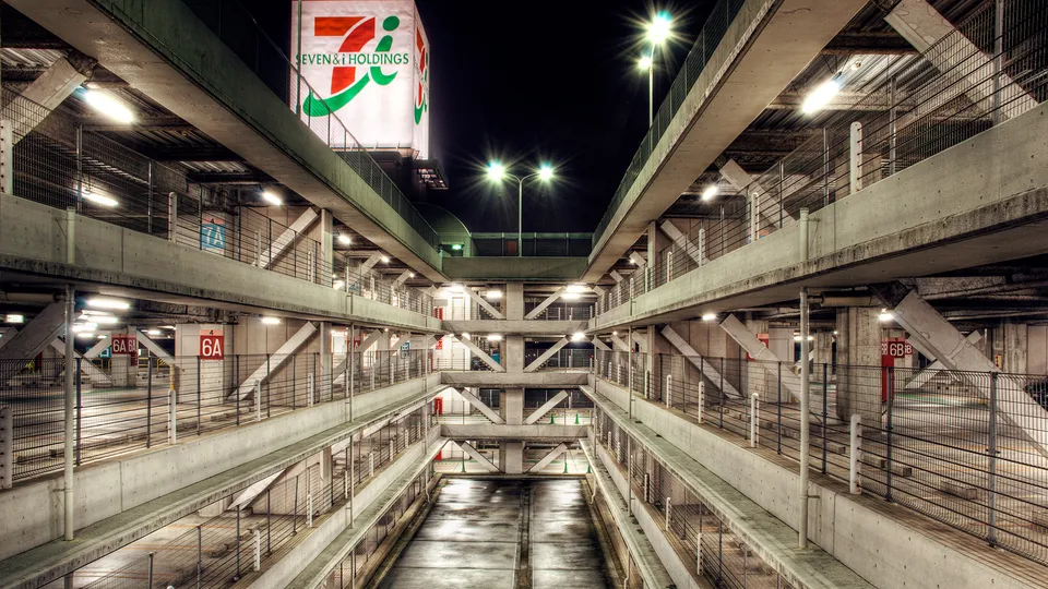 A multi-level parking garage at night, illuminated by artificial lights, with a large Seven & i Holdings sign visible.