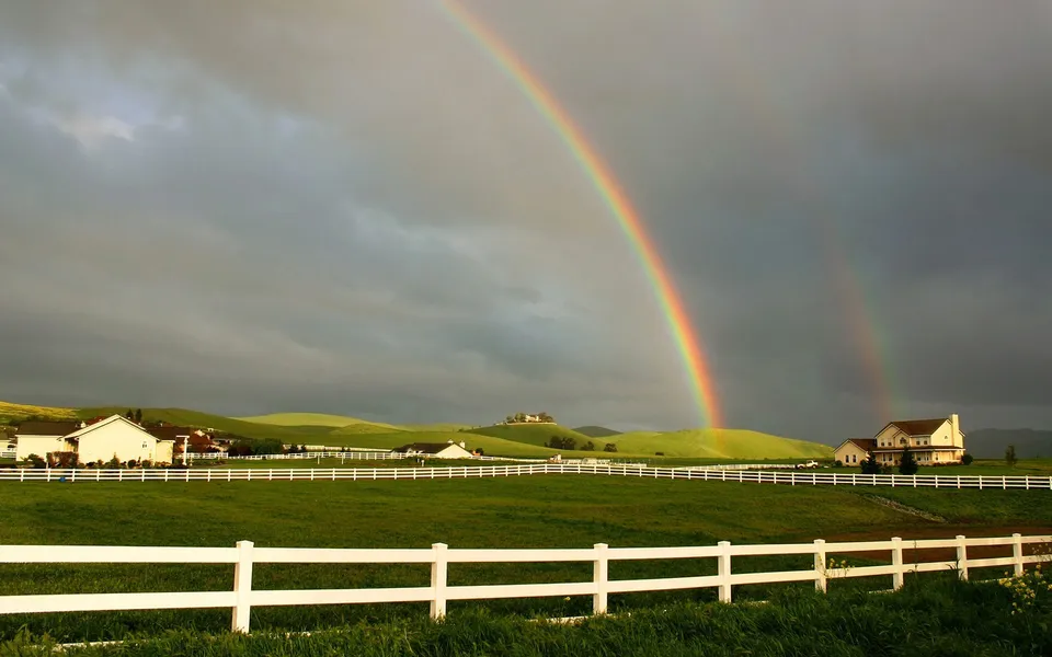 A double rainbow appears over green fields with white fences and houses.
