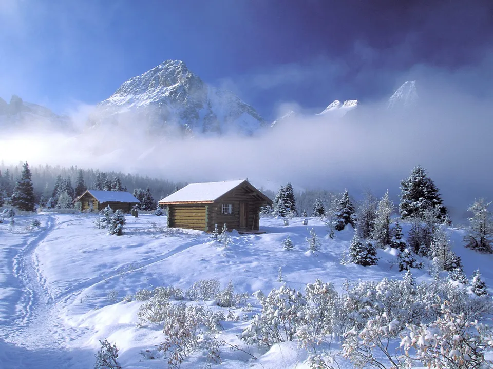 Two log cabins sit in a snowy mountain landscape with mist obscuring the peaks.
