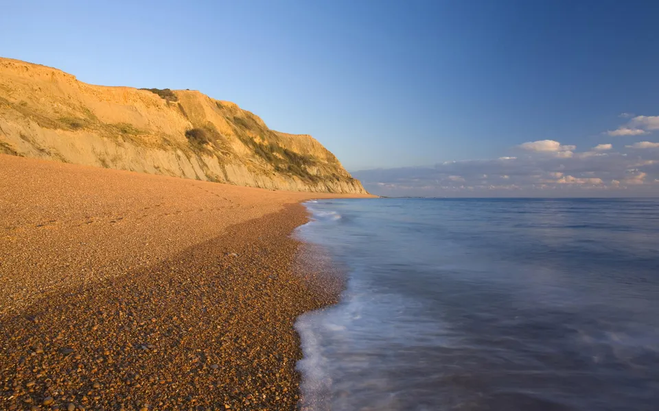 A wide pebble beach stretches along a golden cliff face with gentle waves lapping the shore under a clear blue sky.