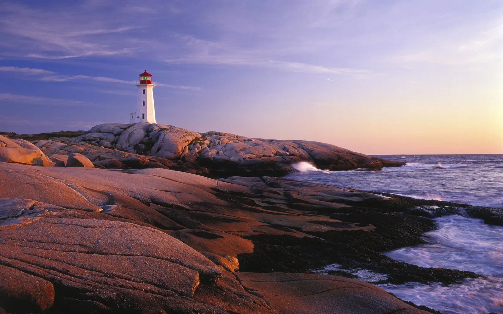 A white lighthouse with a red roof stands on a rocky coastline during sunset.