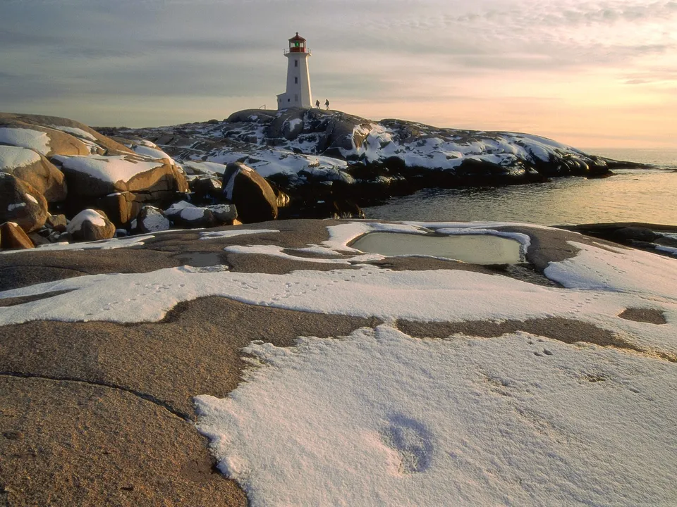 A white lighthouse stands on a rocky outcrop covered in snow, with the ocean in the background.