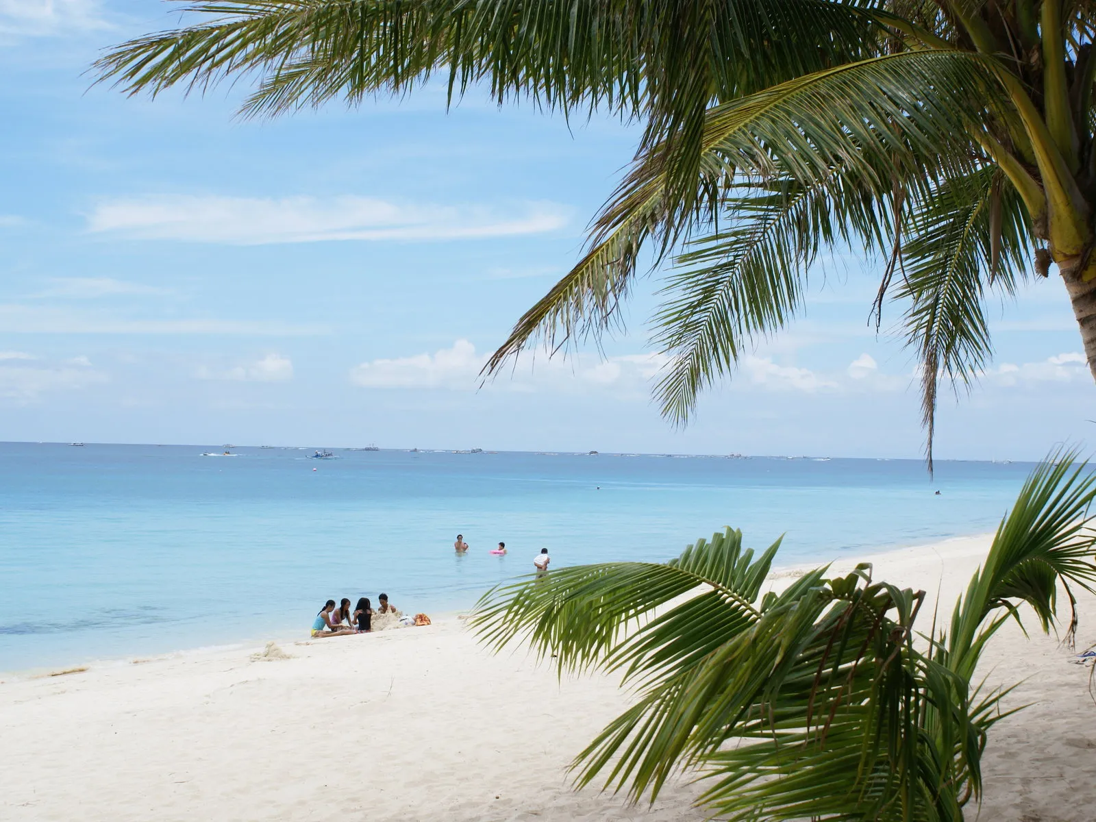 People relax on a white sand beach next to clear turquoise waters with gentle waves lapping the shore under a blue sky with clouds.