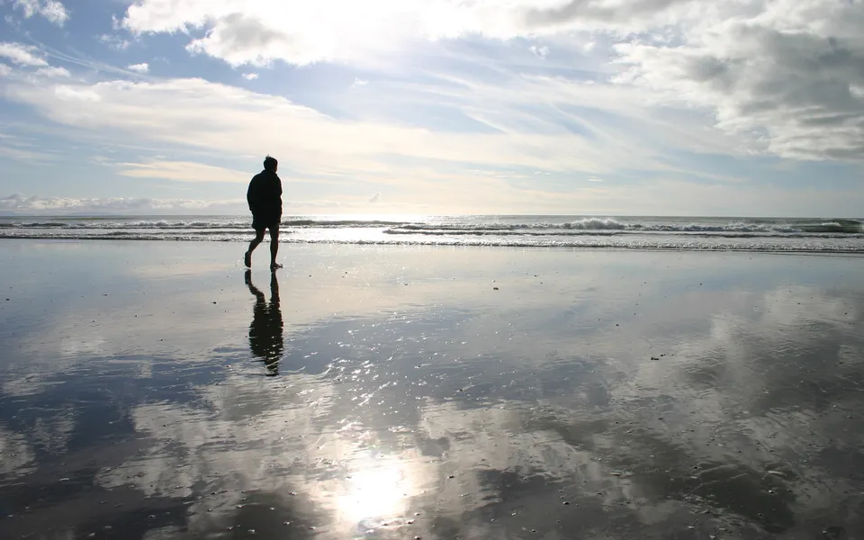 A person walks along a wet beach, their reflection mirrored in the sand under a cloudy sky.