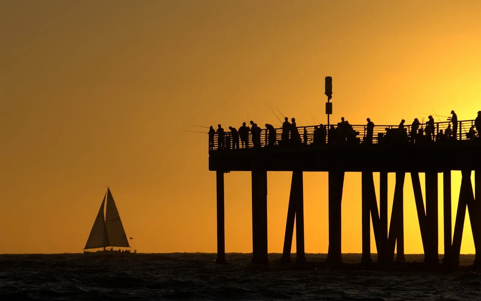 Silhouettes of people fishing on a pier and a sailboat on the ocean during a vibrant sunset.