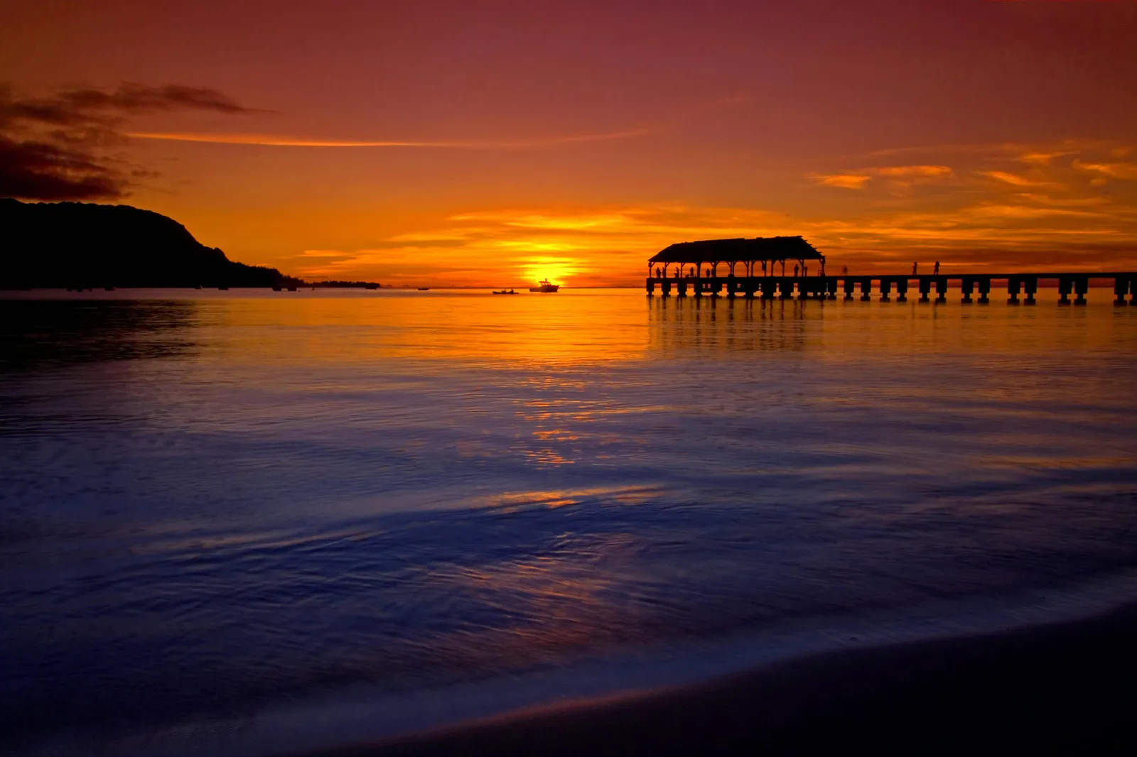A silhouetted pier extends into the ocean during a vibrant sunset with orange and purple skies.