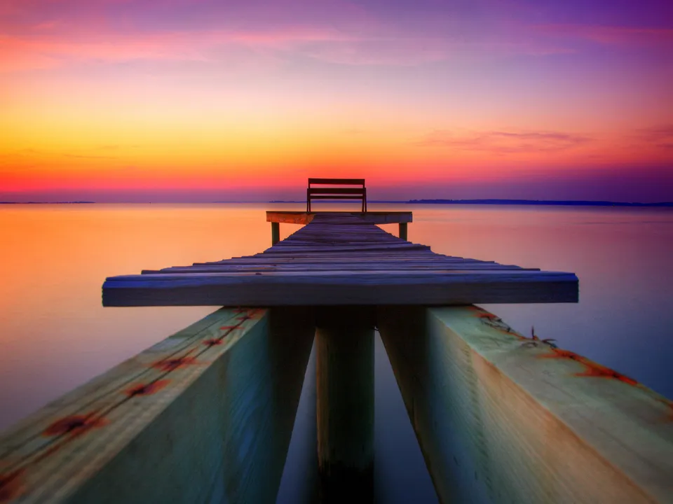 A wooden pier with a bench extends over calm water towards a colorful sunset sky.