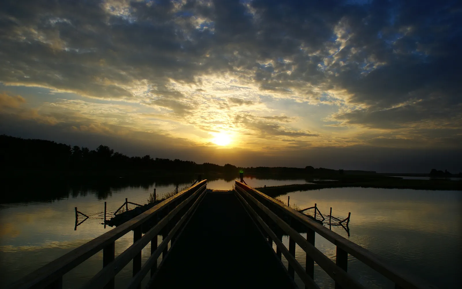 A wooden pier extends over calm water towards a bright sunset with dramatic clouds.