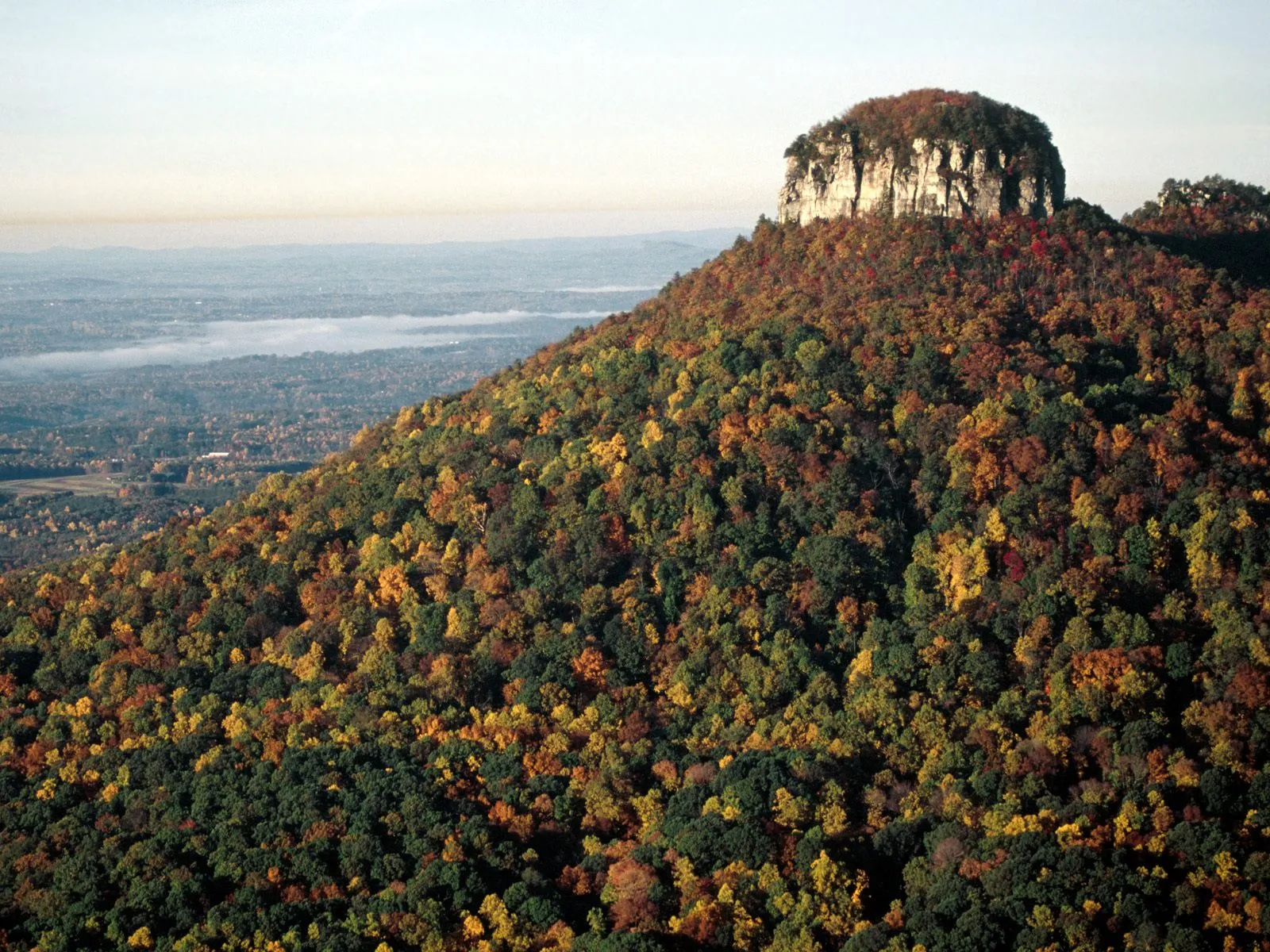 A large, flat-topped mountain covered in colorful autumn trees, with a hazy valley in the distance.