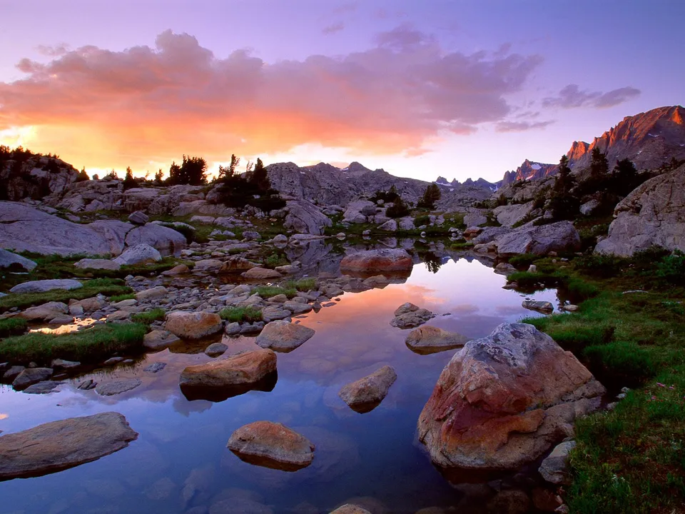 A serene alpine lake reflects a vibrant sunset sky with pink and orange clouds, surrounded by rocky terrain.