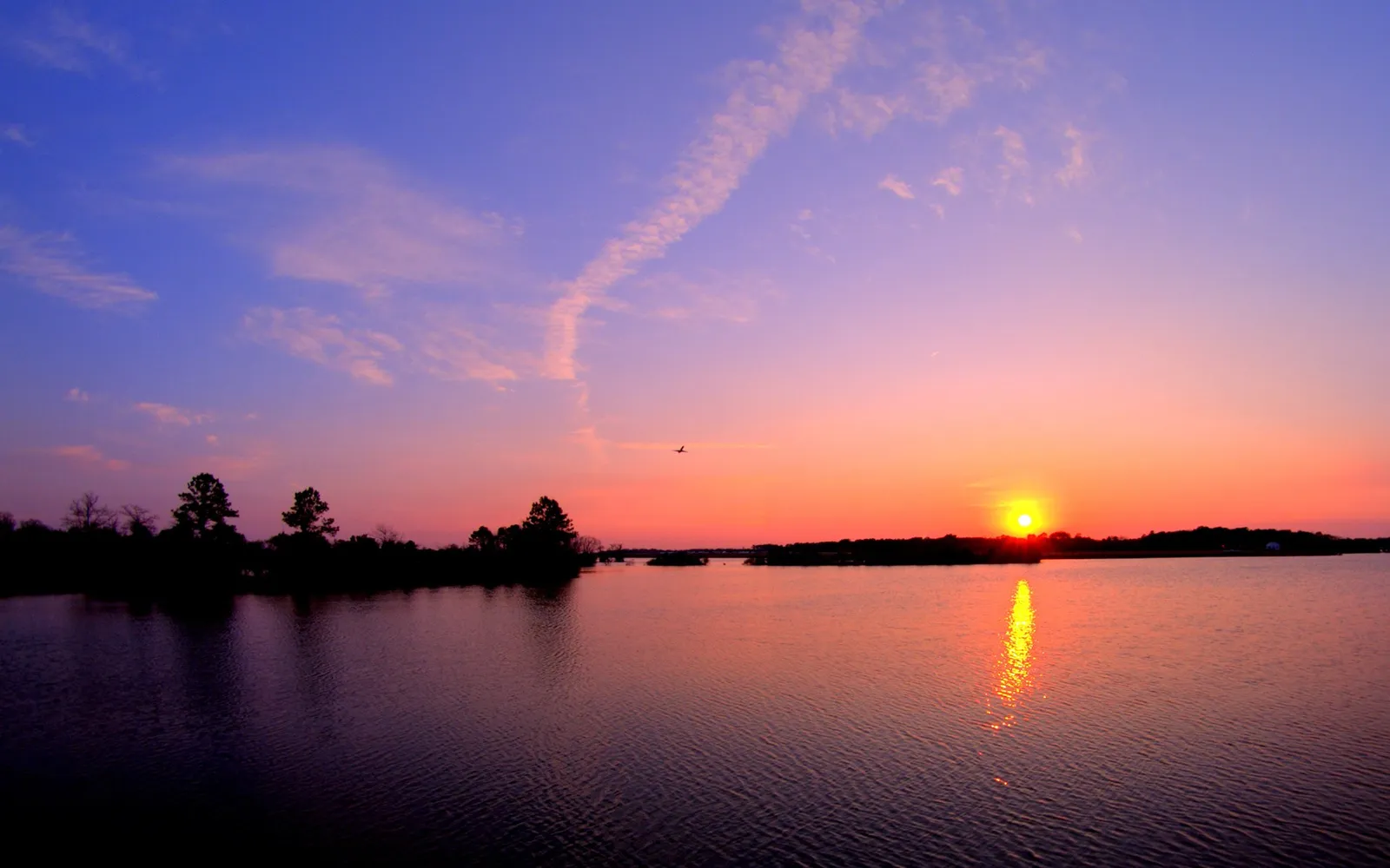 A sunset sky with pink, orange, and purple hues reflects on a tranquil lake with a silhouetted treeline.