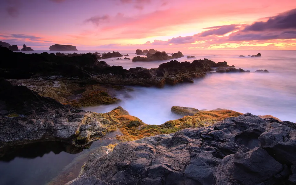 A dramatic sunset paints the sky in shades of pink and purple over a rugged coastline with moss-covered rocks.