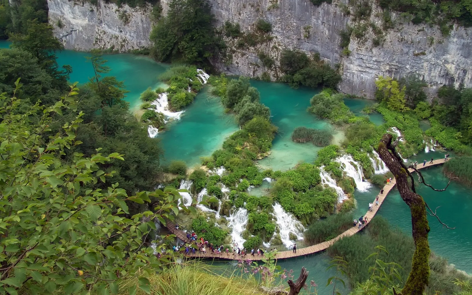 A scenic view of cascading turquoise lakes and waterfalls connected by wooden boardwalks, with lush green vegetation and cliffs.