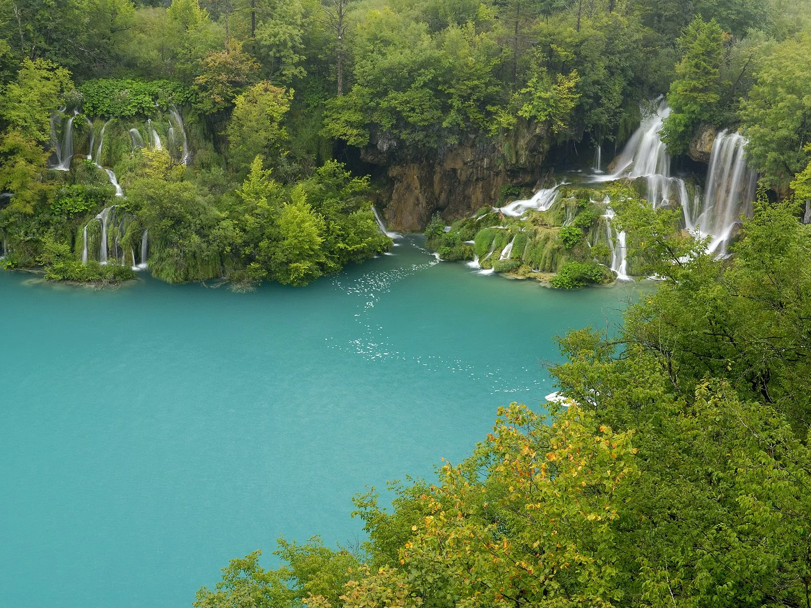 A series of waterfalls cascade down mossy rocks into a vibrant turquoise lake, surrounded by lush green trees.