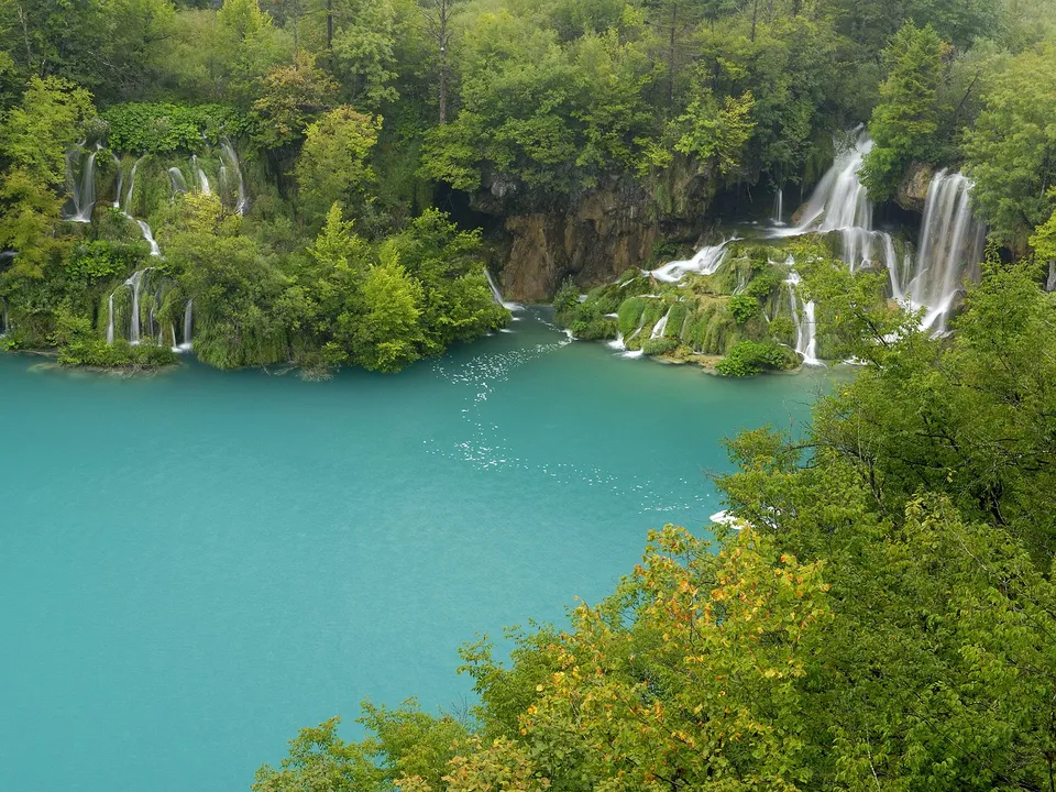 A series of waterfalls cascade down mossy rocks into a vibrant turquoise lake, surrounded by lush green trees.