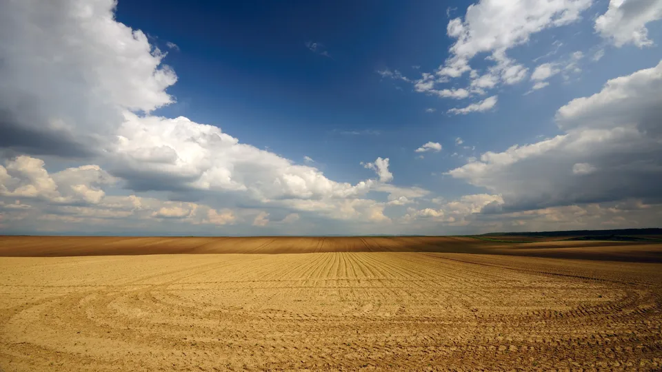 Expansive ploughed fields stretch towards the horizon under a dynamic sky with white clouds and deep blue patches.