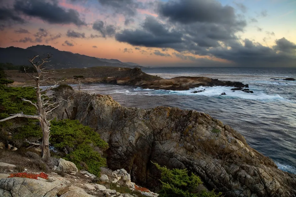 A dramatic sunset with dark clouds over a rocky coastline with crashing waves.