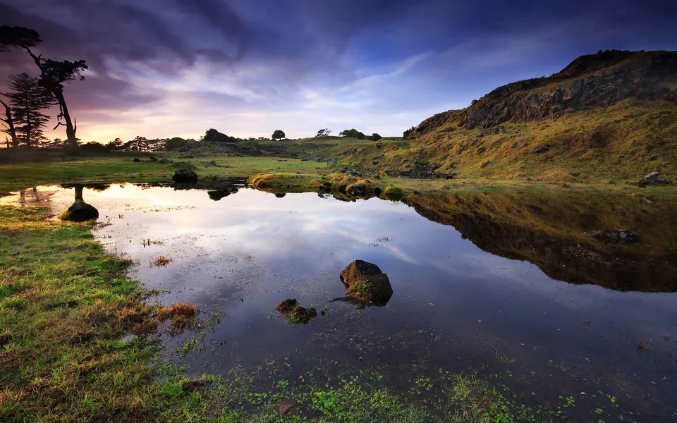 A calm pond reflects a dramatic sky with purple and orange hues during sunset, with grassy hills and trees in the background.