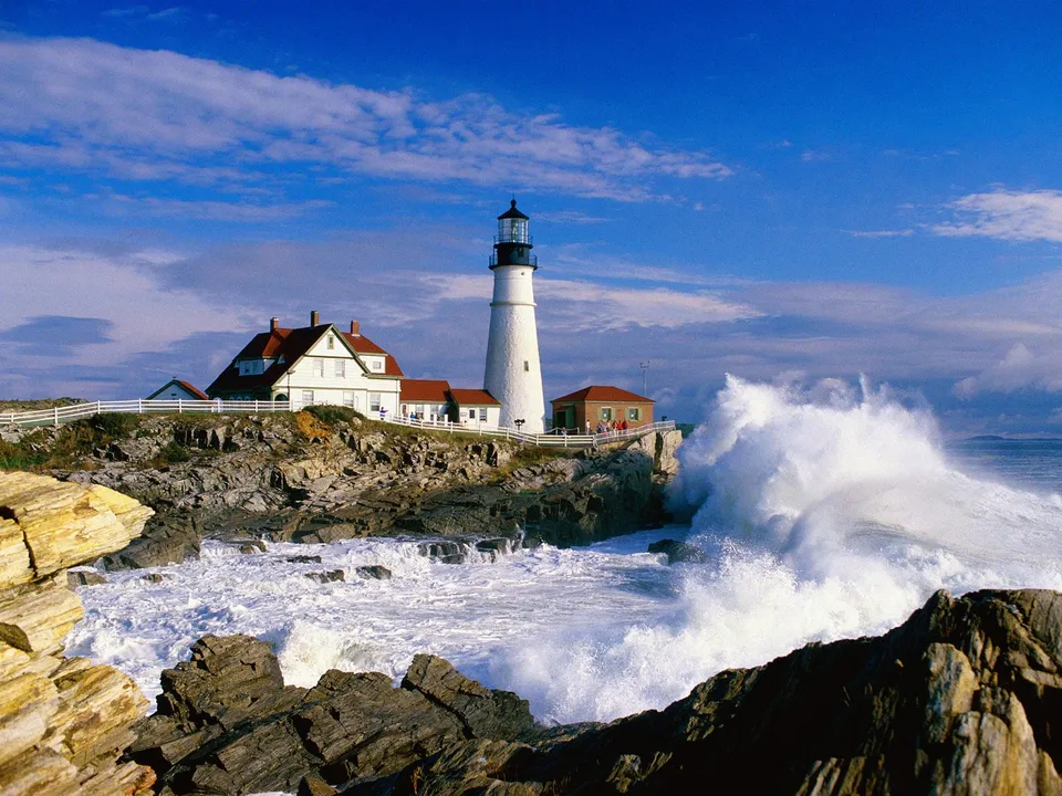 A white lighthouse stands on a rocky coast with large waves crashing against the shore under a blue sky.