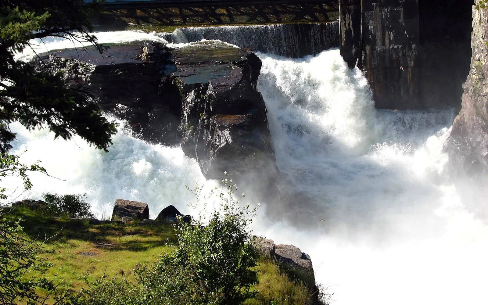 A powerful waterfall cascades over dark, jagged rocks, with mist rising and green grass in the foreground.
